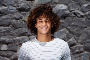 Brown man with big curly hair posing in front of a stone wall smiling
