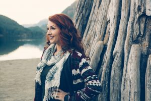 red haired woman wearing a sweater smiling to the distance in front of rocky mountain and lake